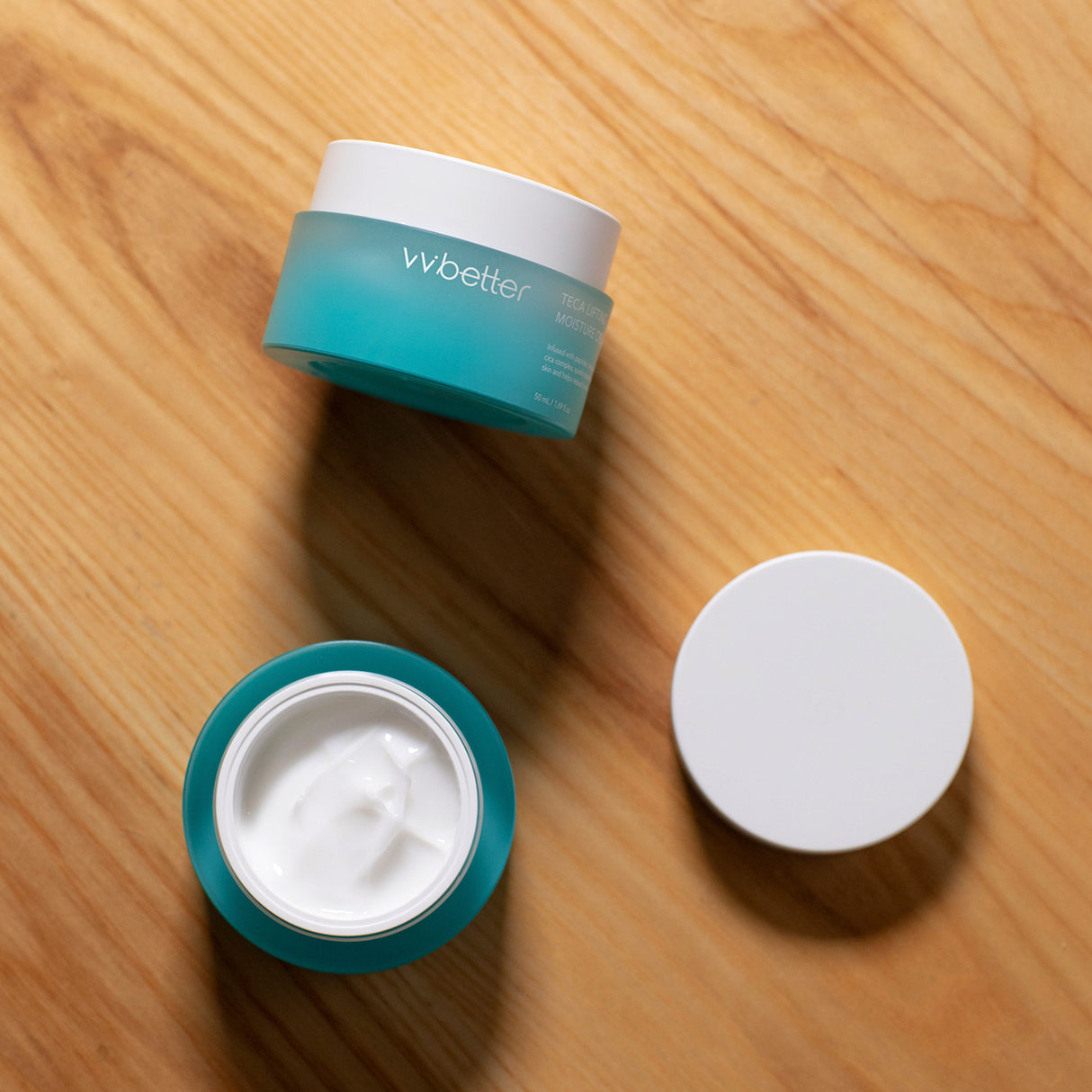 Due vasetti aperti della vvbetter Teca Lifting Moisture Cream su sfondo in legno chiaro, con uno dei barattoli pieno di crema bianca visibile e il coperchio appoggiato accanto, creando un’atmosfera calda e naturale.