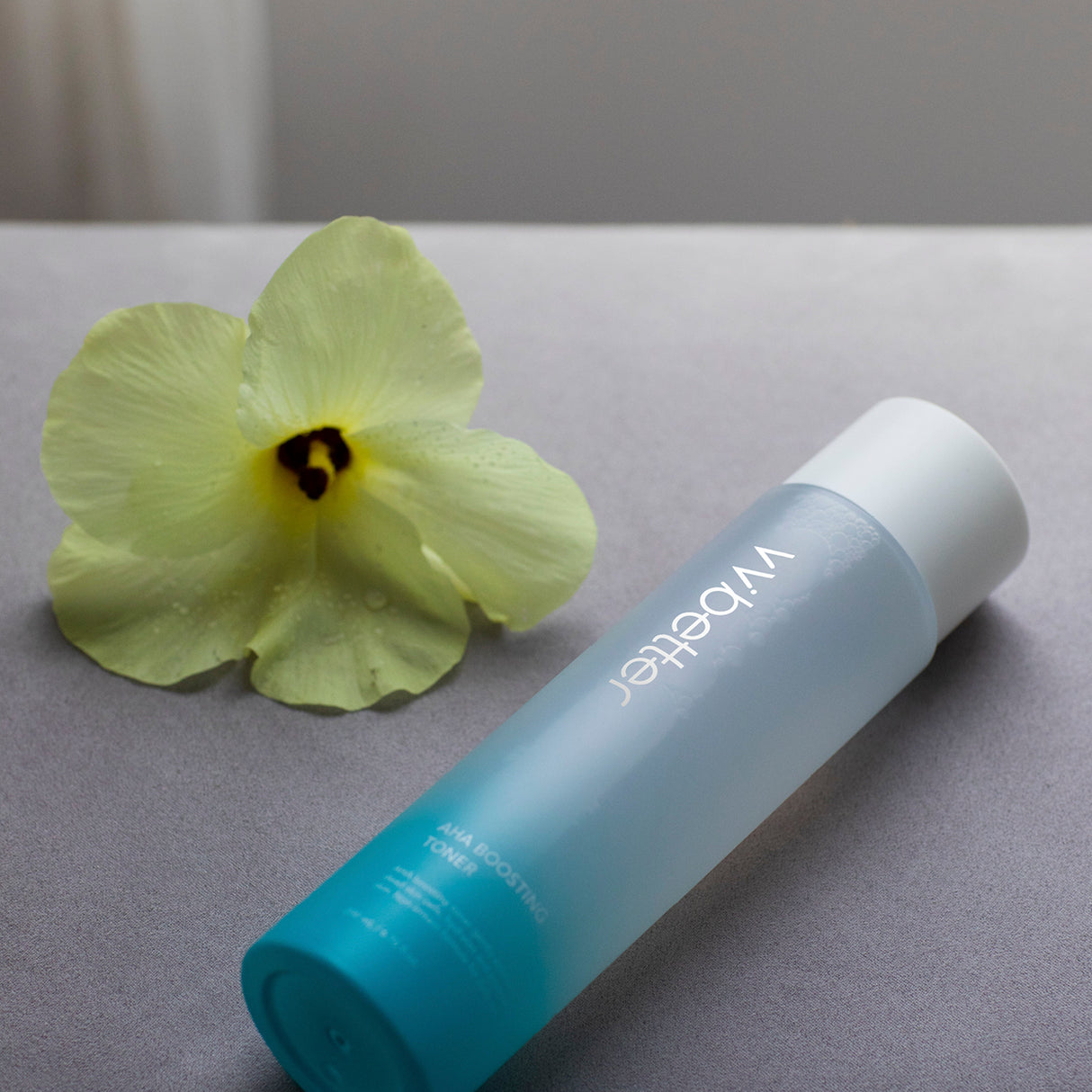 Flacone in vetro satinato sfumato azzurro del tonico "AHA Boosting Toner" di vvbetter, adagiato su una superficie grigia accanto a un fiore giallo chiaro con petali bagnati.
