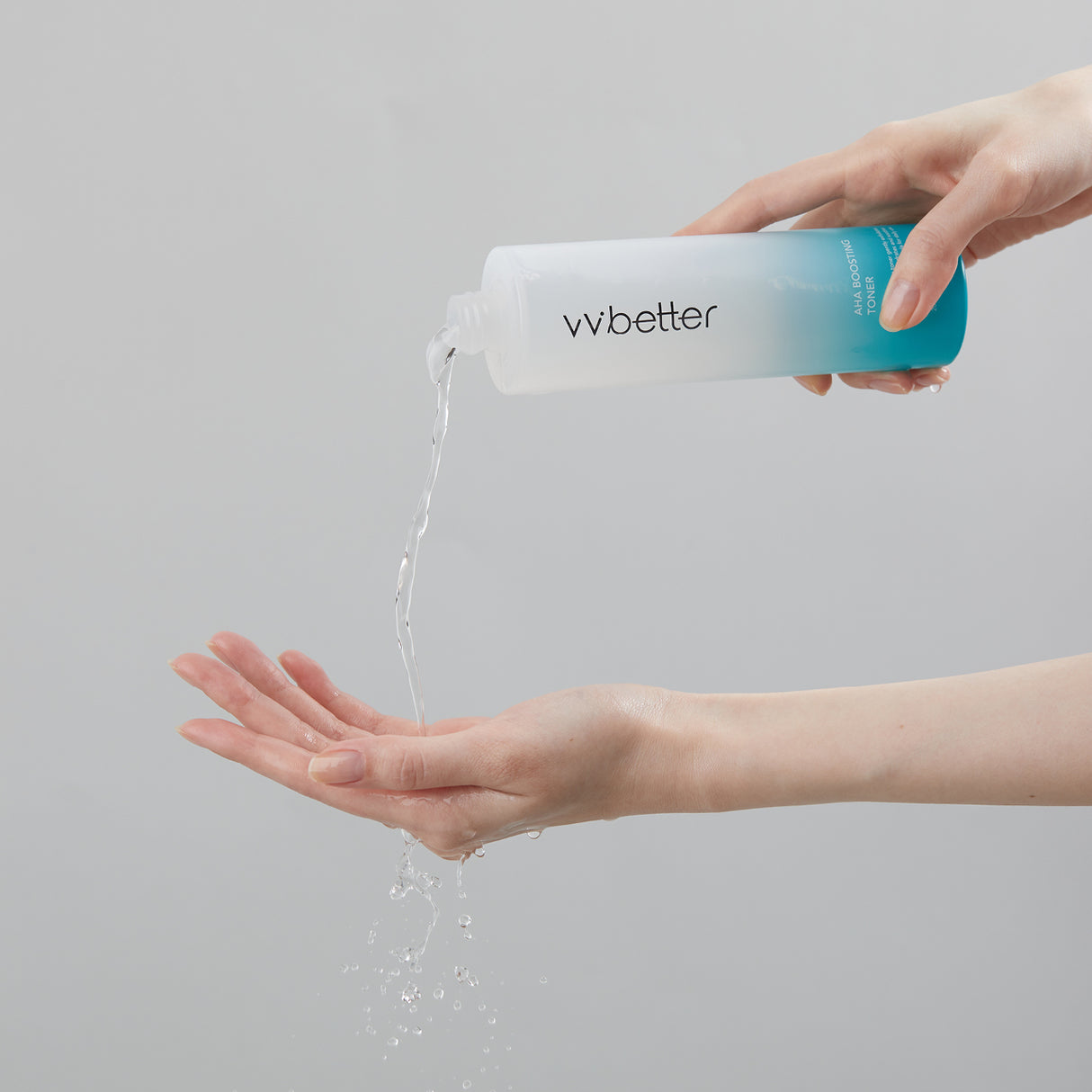 Una mano versa un getto di tonico trasparente da una bottiglia vvbetter AHA Boosting Toner color azzurro sfumato in un’altra mano aperta, su sfondo grigio chiaro.