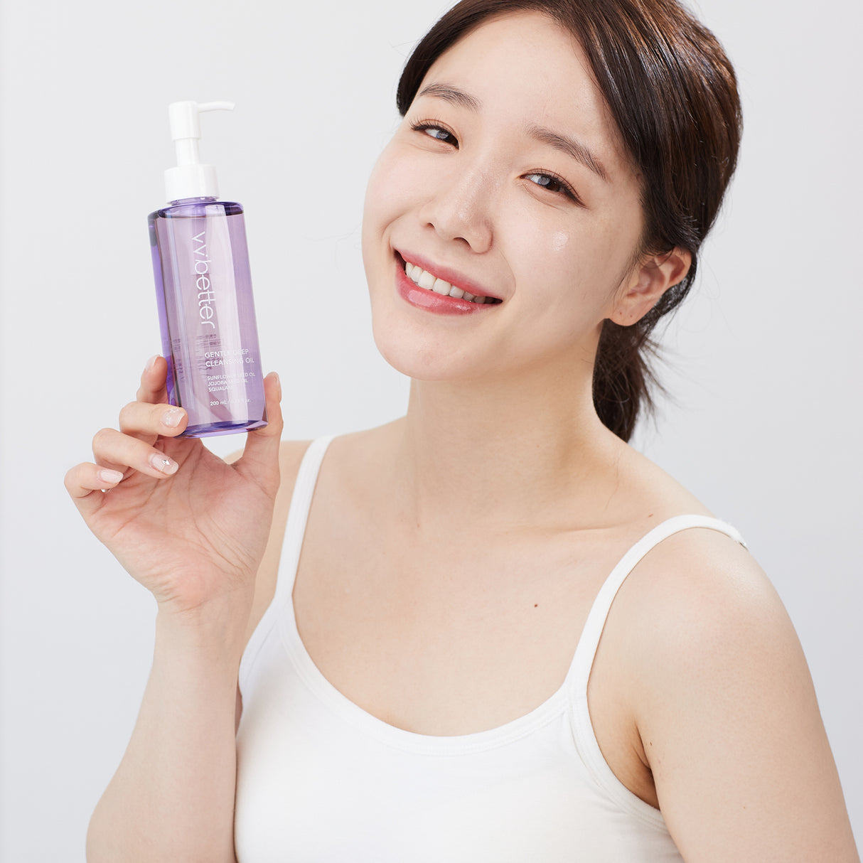 Donna sorridente con top bianco tiene in mano un flacone trasparente color lavanda del vvbetter Gentle Deep Cleansing Oil con dispenser bianco, su sfondo neutro chiaro.