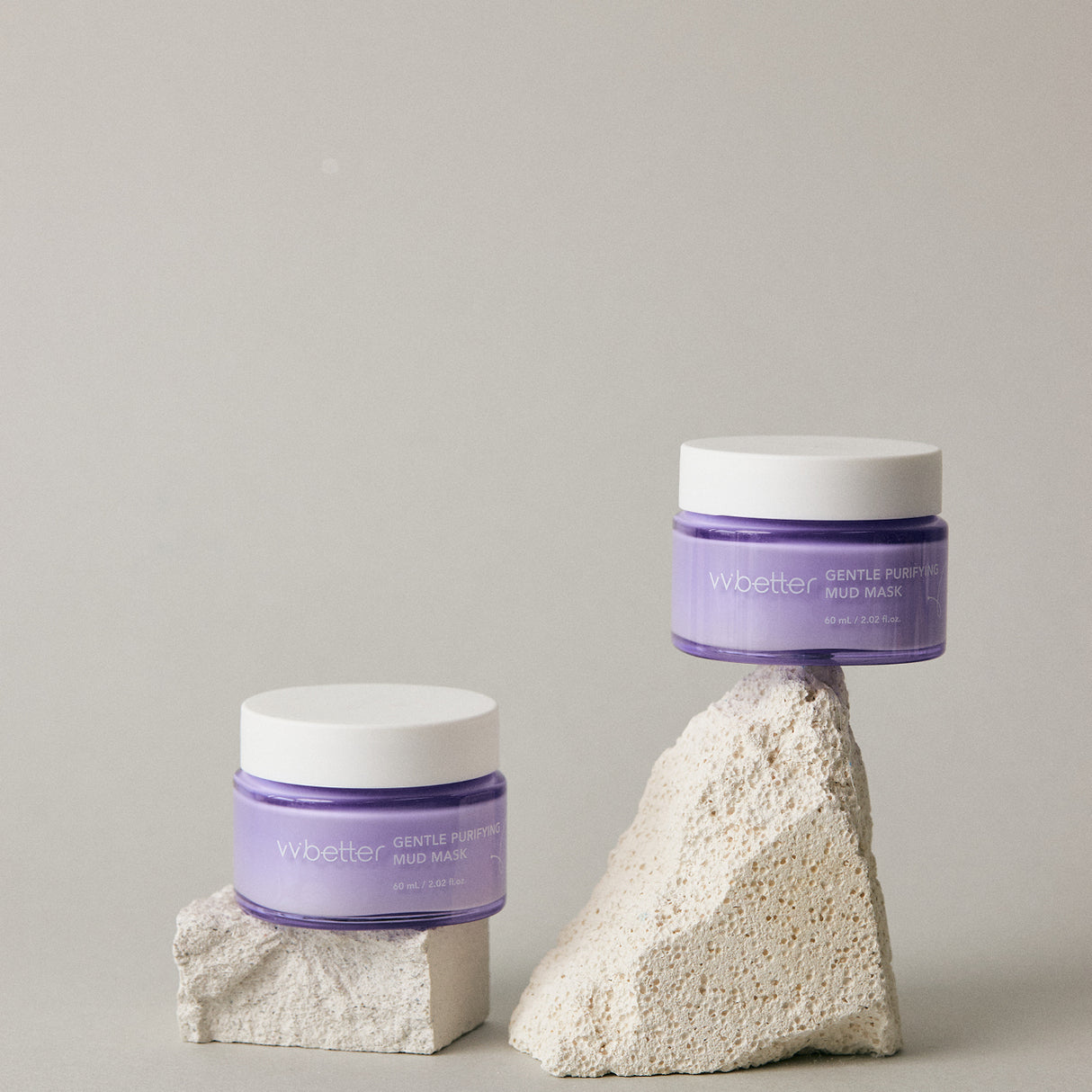 Due vasetti del vvbetter Gentle Purifying Mud Mask in vetro lilla con coperchio bianco, elegantemente posizionati su blocchi di pietra beige. Lo sfondo neutro e minimalista enfatizza il packaging del prodotto, trasmettendo un senso di purezza e cura della pelle.