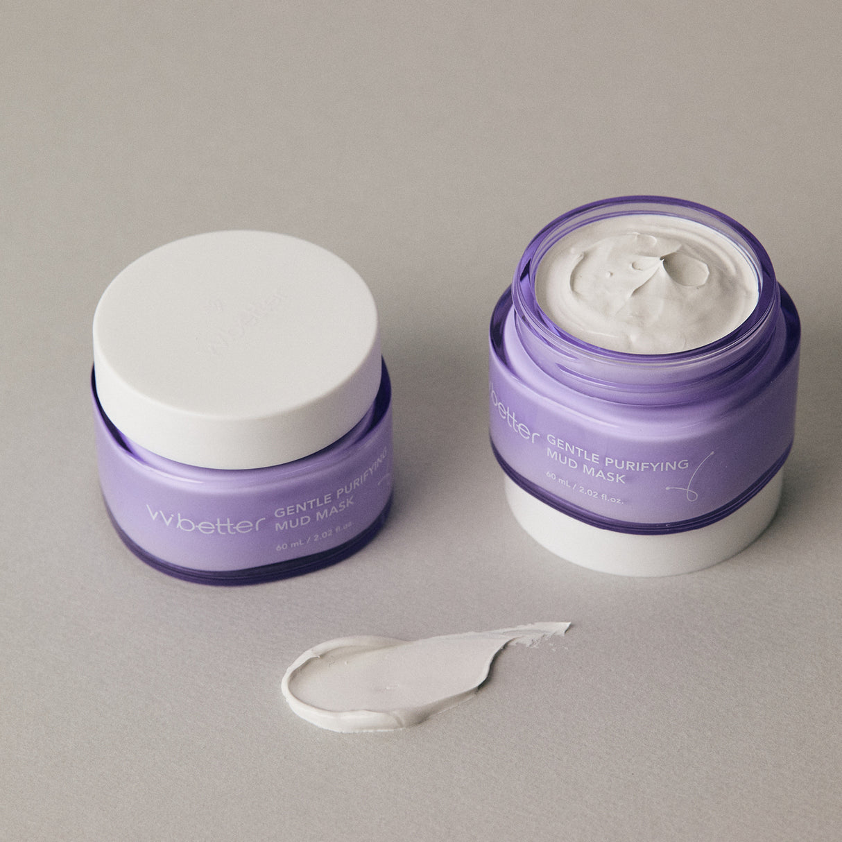 Due barattoli lilla del vvbetter Gentle Purifying Mud Mask, uno chiuso e uno aperto che mostra la texture cremosa della maschera all’interno. Accanto, una piccola quantità di prodotto è stesa elegantemente sulla superficie beige, enfatizzando la consistenza ricca e vellutata del trattamento.