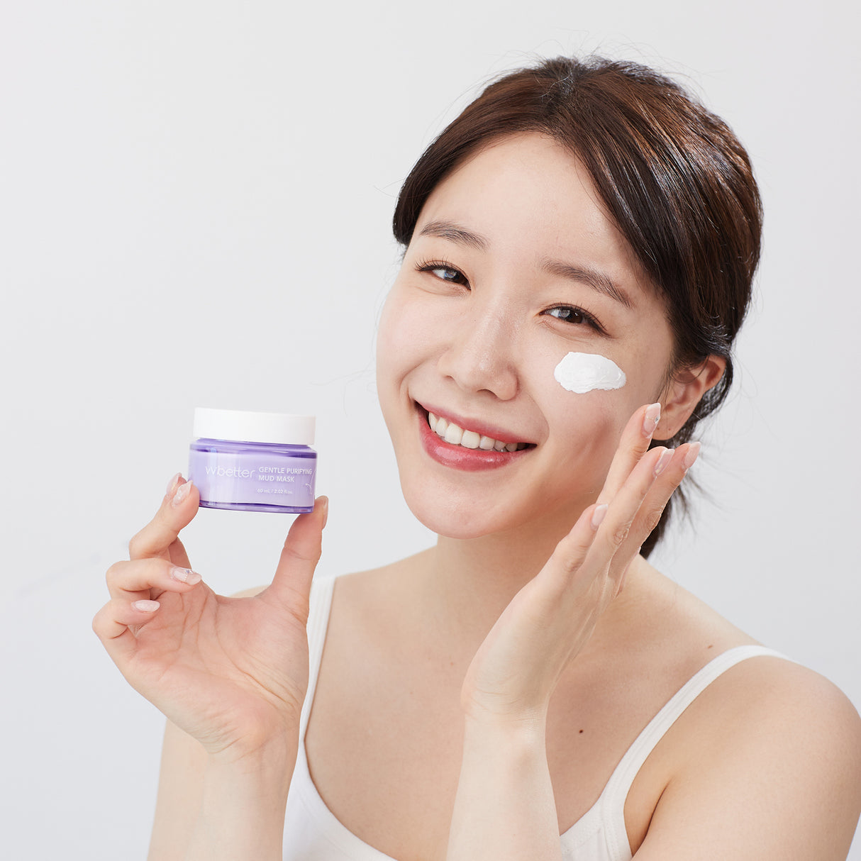 Modella sorridente che applica la vvbetter Gentle Purifying Mud Mask sulla guancia. Tiene in mano il vasetto lilla del prodotto, con una texture cremosa bianca già stesa sul viso. Lo sfondo è bianco, pulito, per enfatizzare la freschezza e la semplicità della skincare.