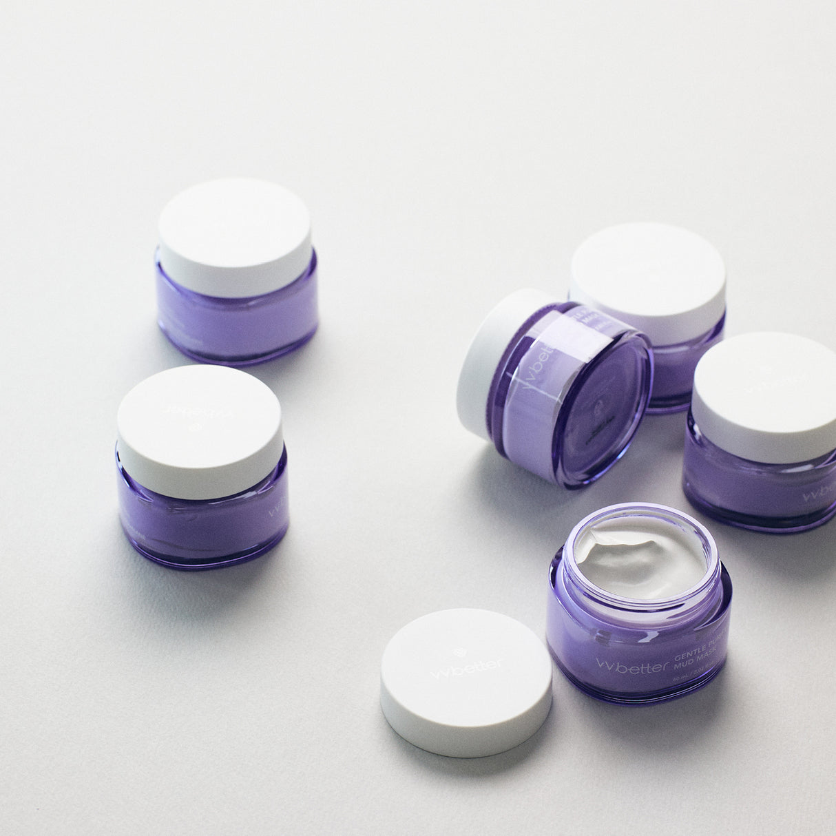 Vasetti lilla di vvbetter Gentle Purifying Mud Mask disposti su una superficie chiara. Alcuni sono chiusi, mentre uno è aperto e mostra la texture cremosa bianca all'interno. L’estetica è pulita, minimal e moderna, in linea con il branding skincare naturale.