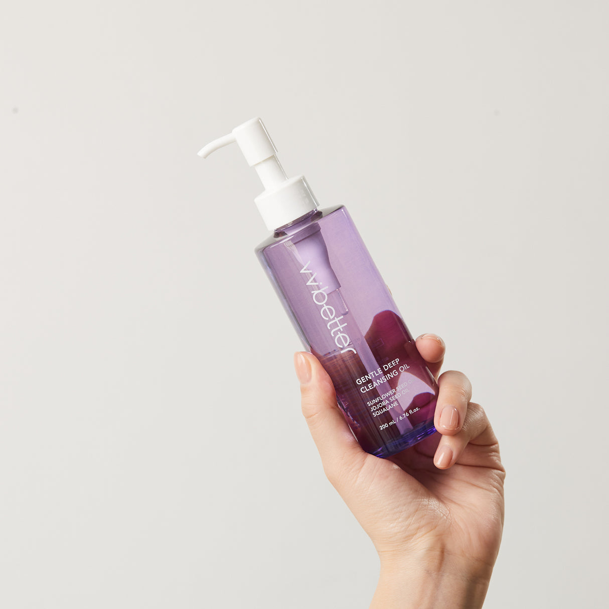 Mano che tiene un flacone trasparente color lavanda del detergente viso vvbetter Gentle Deep Cleansing Oil con dispenser bianco, su sfondo neutro.