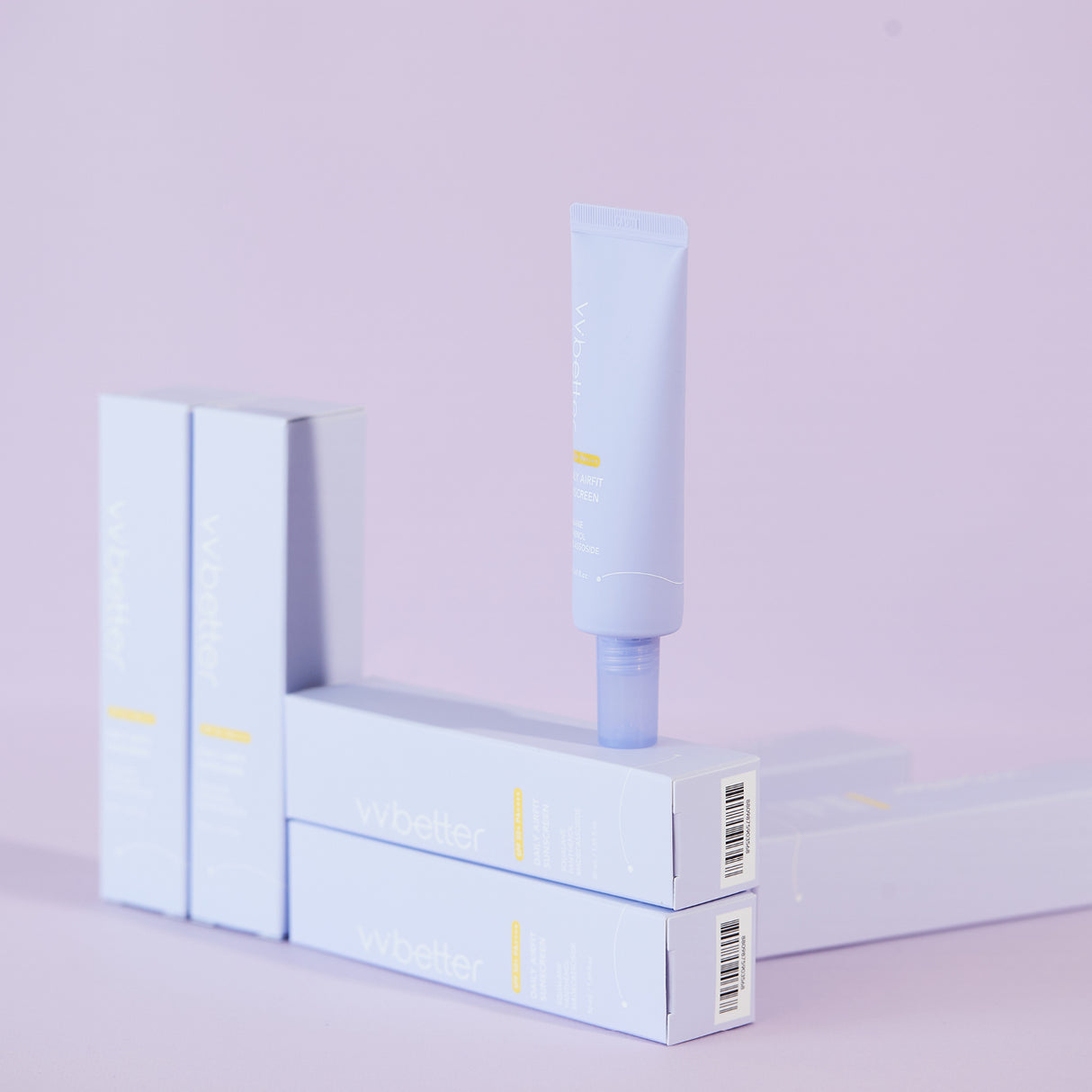 Un tubetto di vvbetter Daily Airfit Sunscreen SPF 50+ PA++++ è posizionato in verticale sopra la sua confezione, con altre scatole allineate in secondo piano. L’intera scena ha uno sfondo lavanda minimalista, creando un effetto visivo pulito ed elegante che richiama la delicatezza del prodotto.