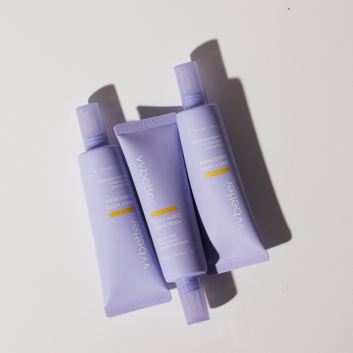Tre tubetti di vvbetter Daily Airfit Sunscreen SPF 50+ PA++++ sono disposti in modo casuale su una superficie bianca. La luce crea ombre nette e profonde, evidenziando le tonalità lavanda opache delle confezioni. Il design minimalista e moderno trasmette freschezza e delicatezza.