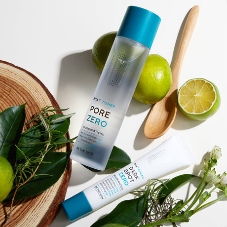 Toner Pore Zero e crema Dark Spot Zero di Be The Skin disposti su una superficie bianca insieme a foglie verdi, lime freschi tagliati e interi, un cucchiaio di legno e un disco di legno naturale, per una composizione ispirata alla natura e alla freschezza.