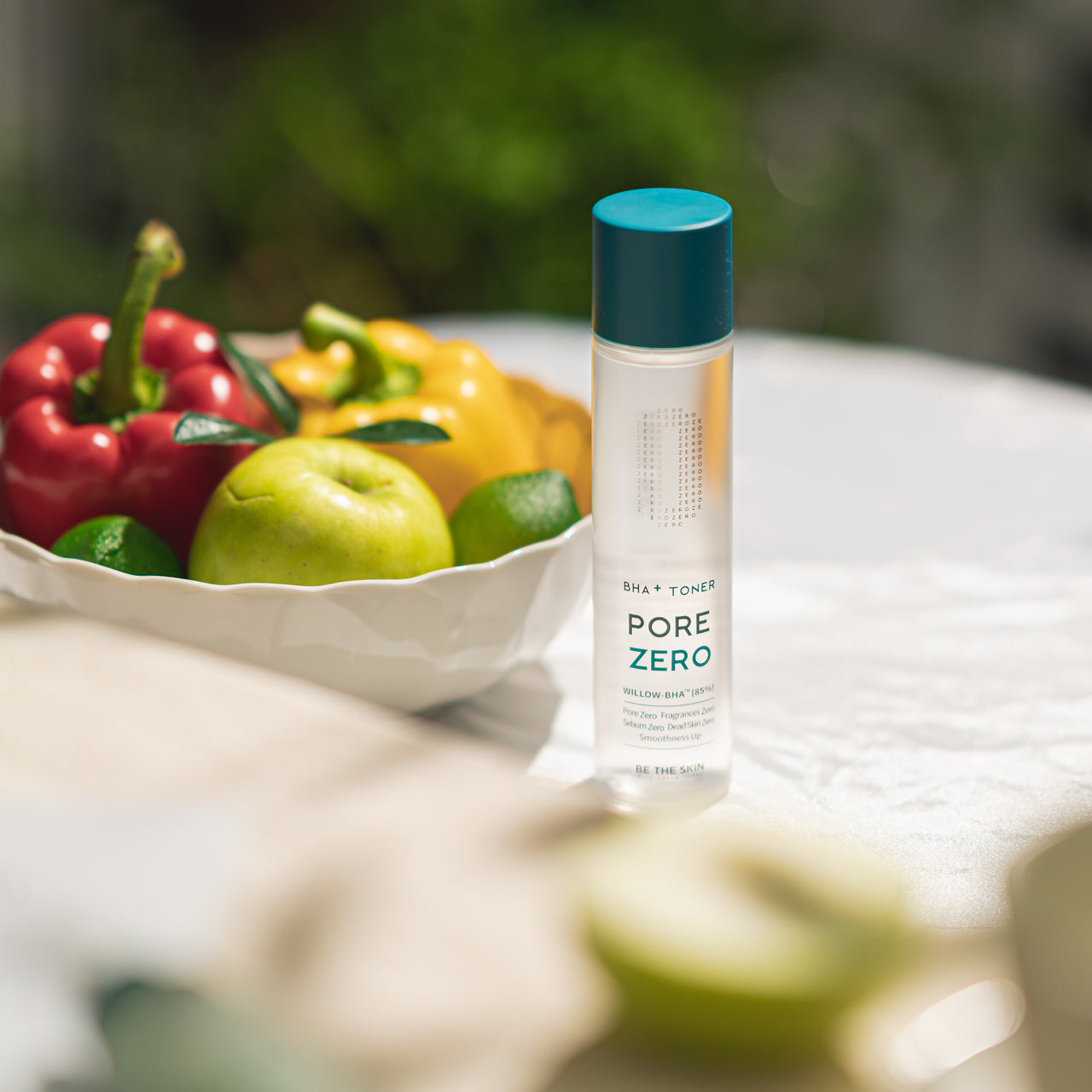 Flacone di toner Pore Zero BHA+ Toner di Be The Skin posizionato su un tavolo accanto a una ciotola con frutta e verdura, tra cui mele verdi, peperoni rossi e gialli, e lime, in un ambiente esterno soleggiato.