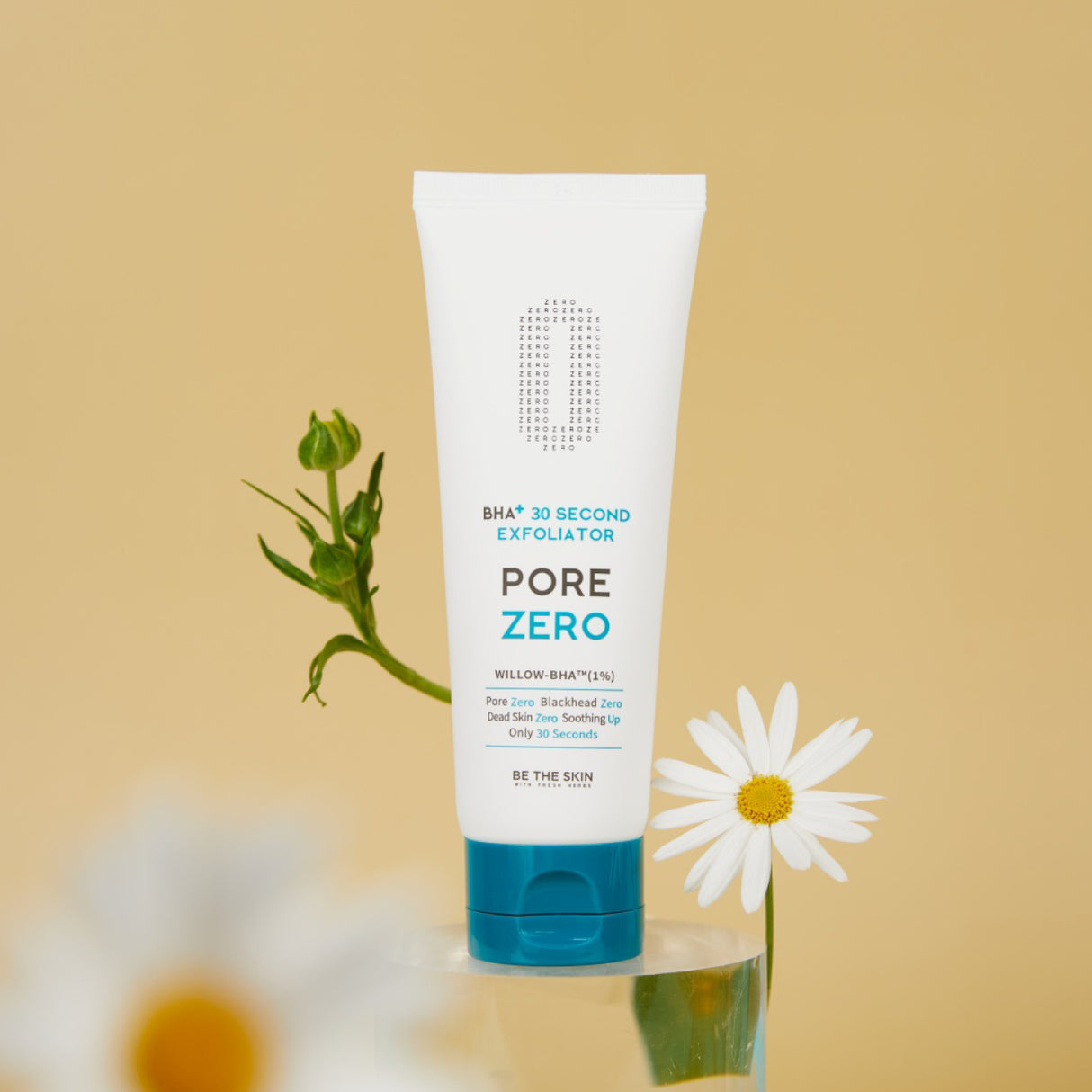 Tubo dell’esfoliante BE THE SKIN PORE ZERO BHA+ 30 Second Exfoliator in piedi su una superficie trasparente, affiancato da margherite bianche, su sfondo beige chiaro.

