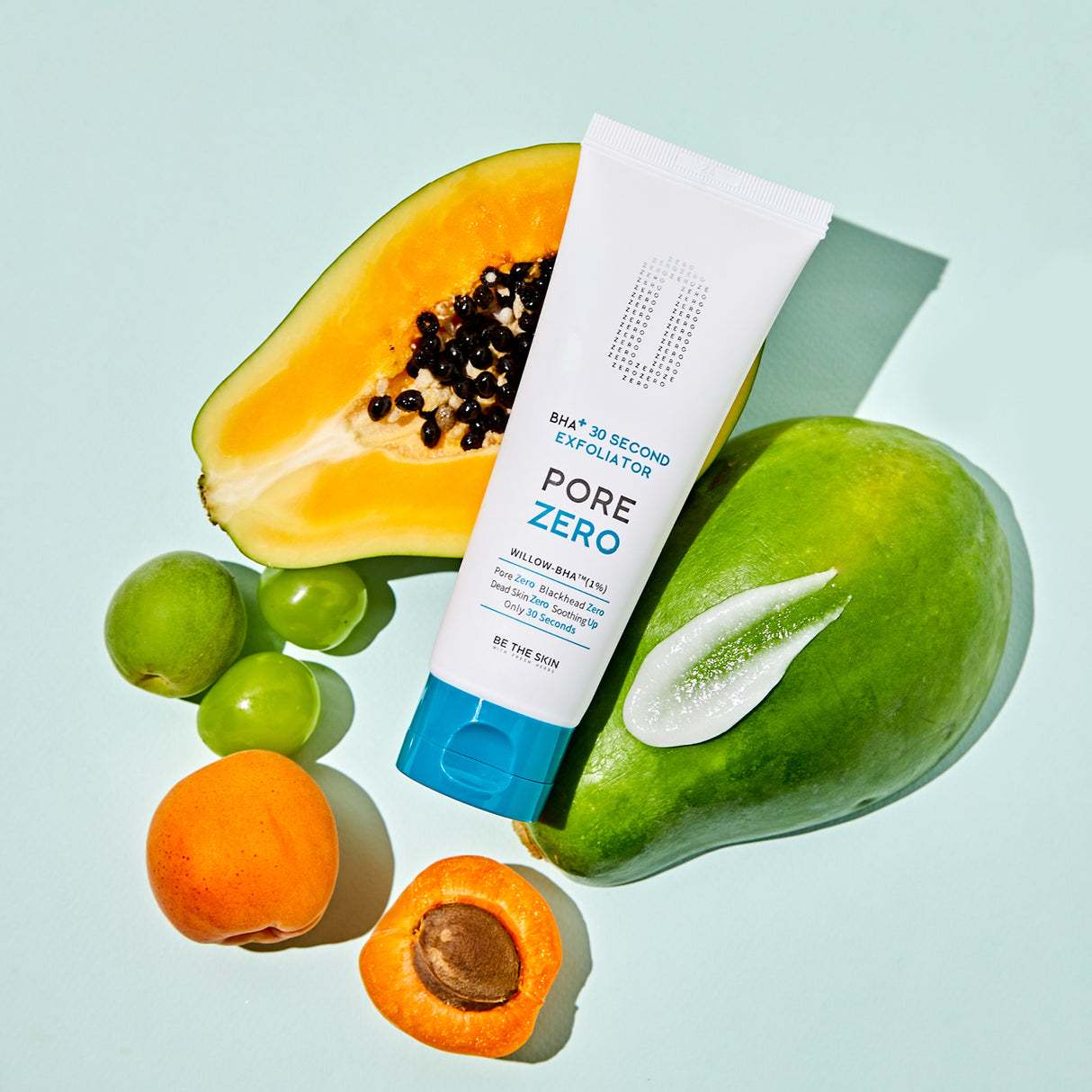 Tubo dell’esfoliante BE THE SKIN PORE ZERO BHA+ 30 Second Exfoliator adagiato tra frutta fresca tropicale, inclusi papaya, mango, albicocche e uva verde, con una texture di prodotto visibile sopra il mango, su sfondo verde chiaro.