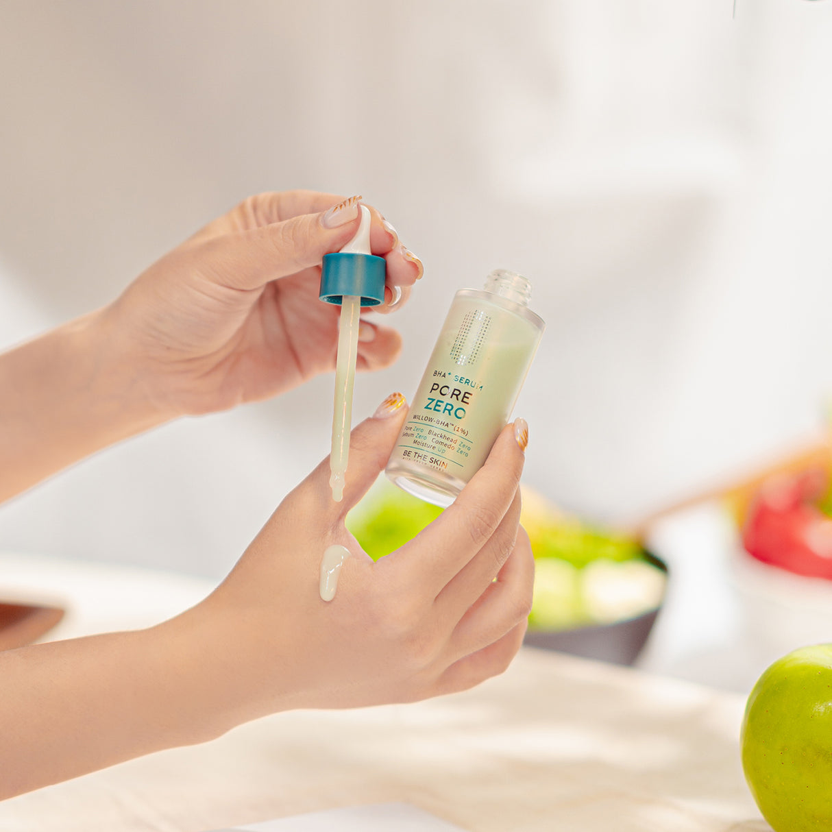 Mani che applicano il siero BE THE SKIN PORE ZERO BHA+ con contagocce, facendo cadere alcune gocce sulla pelle della mano, con sfondo sfocato e luminoso.

