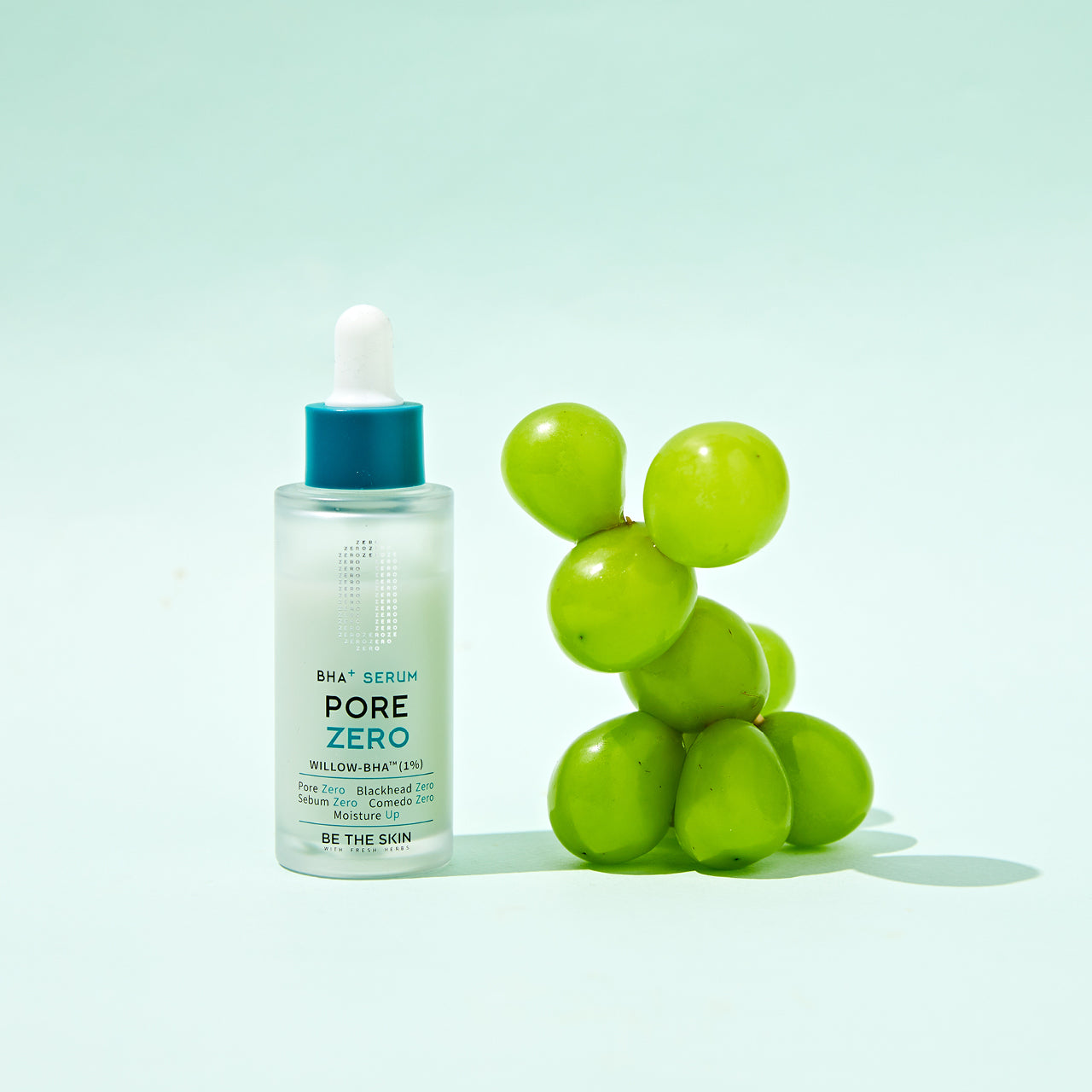 Flacone del siero BE THE SKIN PORE ZERO BHA+ con contagocce, posizionato accanto a un grappolo di uva verde su sfondo verde pastello, evocando freschezza e ingredienti naturali.