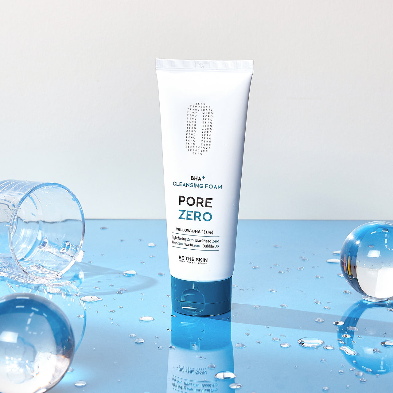 Detergente viso BE THE SKIN PORE ZERO BHA+ CLEANSING FOAM in piedi su superficie blu riflettente, con gocce d’acqua e bicchieri capovolti intorno.

