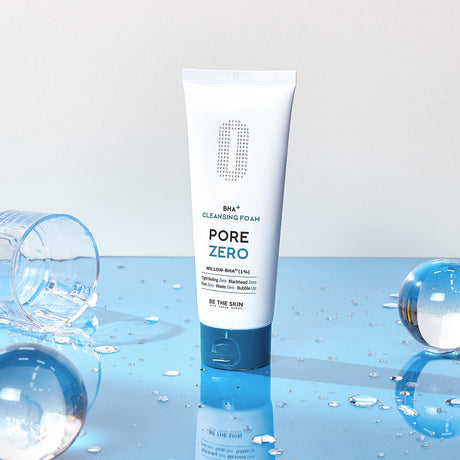Detergente viso BE THE SKIN PORE ZERO BHA+ CLEANSING FOAM in piedi su superficie blu riflettente, con gocce d’acqua e bicchieri capovolti intorno.

