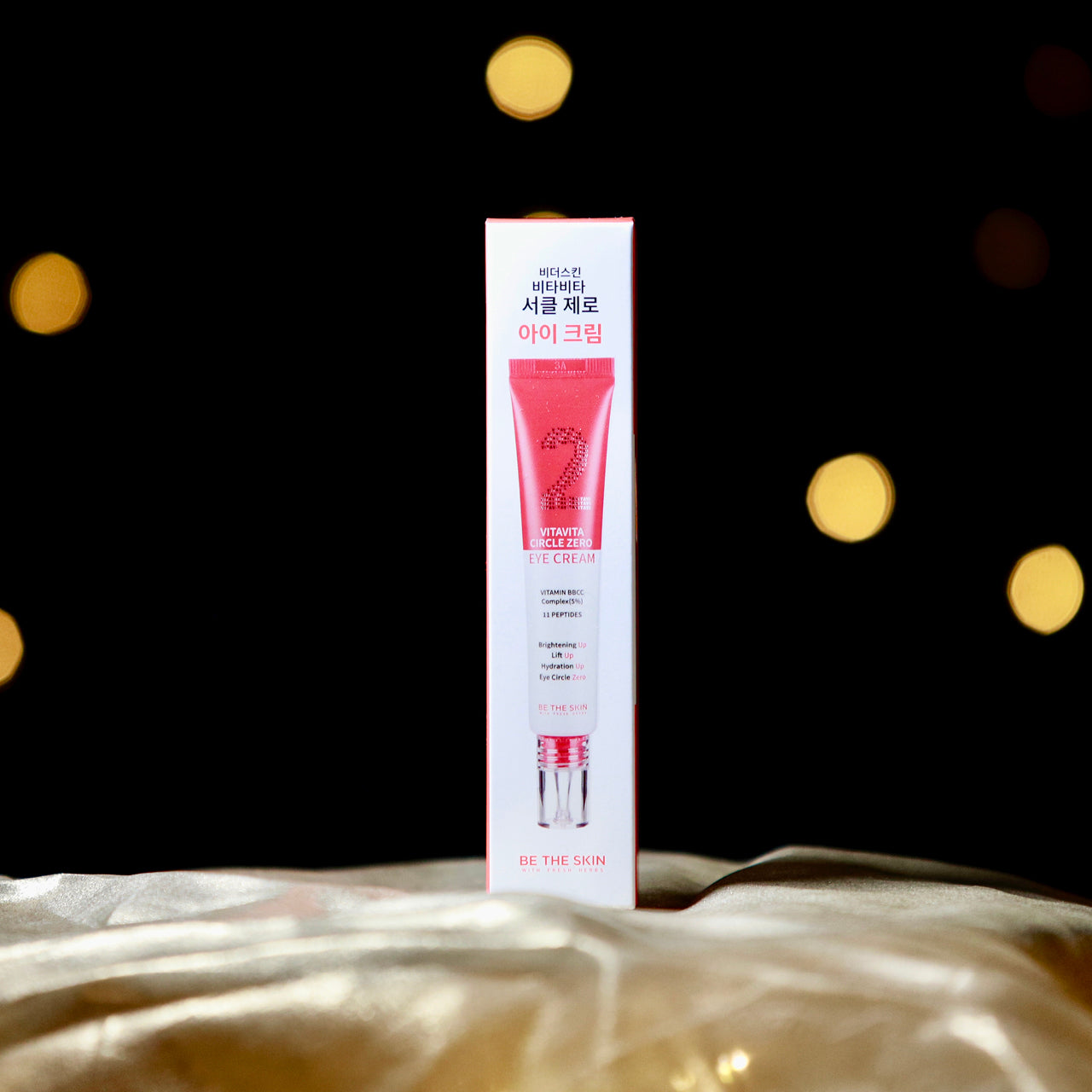 
Scatola della crema contorno occhi "Vitavita Circle Zero Eye Cream" di Be The Skin, posizionata su un tessuto dorato con sfondo nero decorato da luci soffuse e sfocate. Il packaging rosso e bianco evidenzia ingredienti come il complesso Vitamin BBCC (5%) e 11 peptidi, con benefici dichiarati come illuminazione, effetto lift, idratazione e riduzione delle occhiaie.


