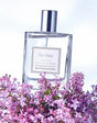Flacone di profumo Like Lilac Eau de Toilette 50 ml in vetro trasparente con tappo argentato, fotografato su sfondo chiaro con fiori di lillà viola in primo piano.
