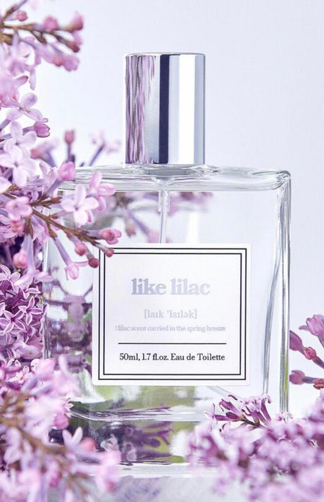 Flacone di profumo coreano Like Lilac Eau de Toilette da 50 ml circondato da fiori di lillà viola su sfondo chiaro, con packaging minimal in vetro trasparente e tappo argentato.
