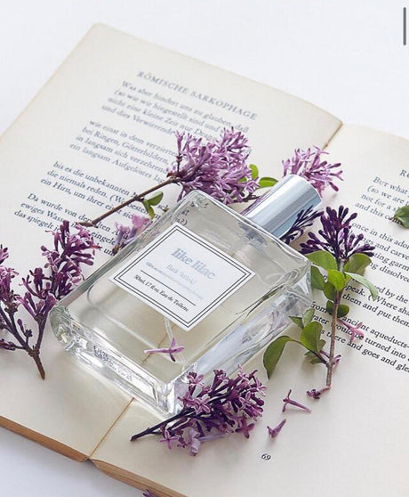Flacone di profumo coreano Like Lilac Eau de Toilette appoggiato su un libro aperto e circondato da fiori di lillà viola, con packaging in vetro trasparente e tappo argentato.
