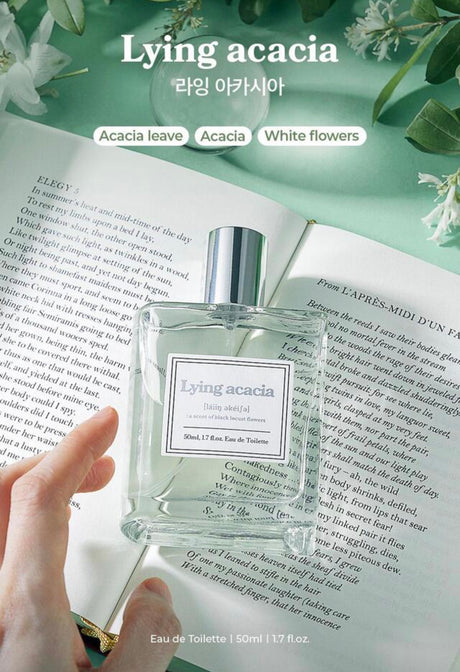Flacone del profumo coreano Lying Acacia appoggiato su un libro aperto, circondato da foglie e fiori bianchi, con indicazione delle note olfattive di acacia, foglie verdi e fiori bianchi.