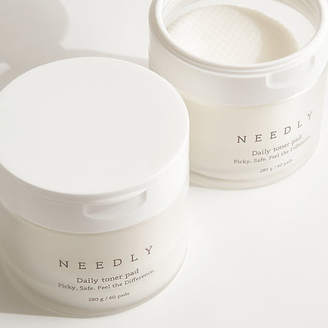 Due barattoli del "Needly Daily Toner Pad" su sfondo chiaro: uno chiuso in primo piano e uno aperto sullo sfondo con i dischetti tonificanti visibili all'interno, evidenziando la texture a rilievo.