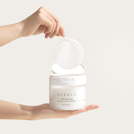Una mano tiene un barattolo aperto di "Needly Daily Toner Pad", mentre un'altra mano solleva delicatamente un dischetto tonificante con pinzetta, mettendo in evidenza la texture a nido d'ape del pad su sfondo neutro.