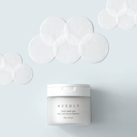 Vasetto del prodotto "NEEDLY Daily Toner Pad" in primo piano su sfondo azzurro chiaro, circondato da numerosi dischetti tonificanti bianchi con texture a nido d'ape, disposti artisticamente intorno.