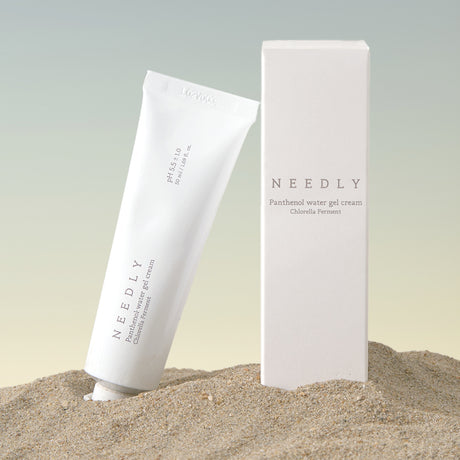 Il tubetto e la confezione della Needly Panthenol Water Gel Cream con Chlorella Ferment sono posizionati nella sabbia su sfondo sfumato beige-azzurro, creando un’atmosfera naturale e rilassante.