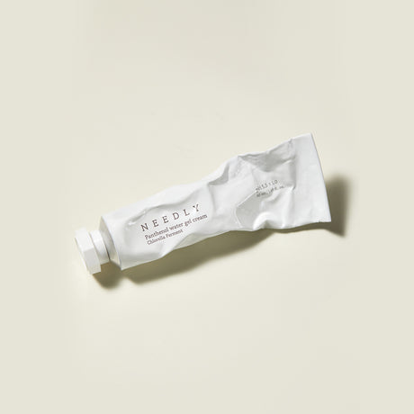 Il tubo bianco leggermente schiacciato della Needly Panthenol Water Gel Cream è appoggiato su una superficie beige chiaro, con tappo chiuso e ombra laterale visibile.

