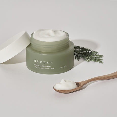 Crema viso Needly Cicachid Relief Cream 48 ml con Chamaecyparis Obtusa Water, texture ricca visibile nel vasetto aperto, con cucchiaino in legno e foglia decorativa su sfondo bianco.