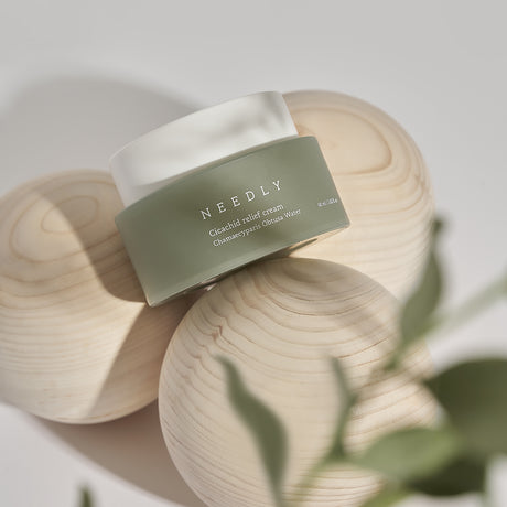 Crema viso Needly Cicachid Relief Cream 48 ml con Chamaecyparis Obtusa Water, confezione verde con tappo bianco, appoggiata su sfere di legno chiaro con decorazioni naturali in primo piano.