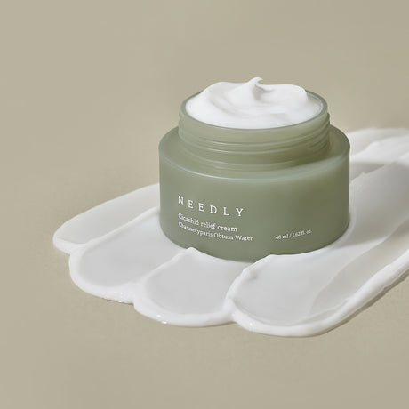 Crema viso Needly Cicachid Relief Cream 48 ml con Chamaecyparis Obtusa Water, vasetto verde su sfondo beige con texture del prodotto spalmata artisticamente alla base.

