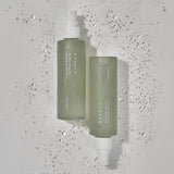 Due flaconi verdi opachi della Needly Cicachid Relaxing Mist adagiati su una superficie bianca bagnata, con gocce d’acqua che evocano freschezza e idratazione.

