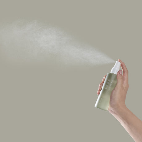 Mano che spruzza la Needly Cicachid Relaxing Mist, mostrando una nuvola sottile e diffusa di prodotto su sfondo beige, enfatizzando l'effetto rinfrescante e leggero dello spray viso.

