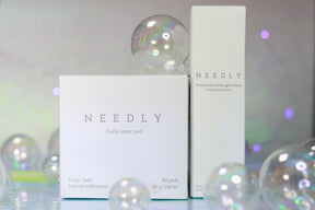 Due confezioni dei prodotti Needly — il Daily Toner Pad e la Panthenol Water Gel Cream — su sfondo perlato con bolle trasparenti decorative, in un’ambientazione luminosa e delicata.

