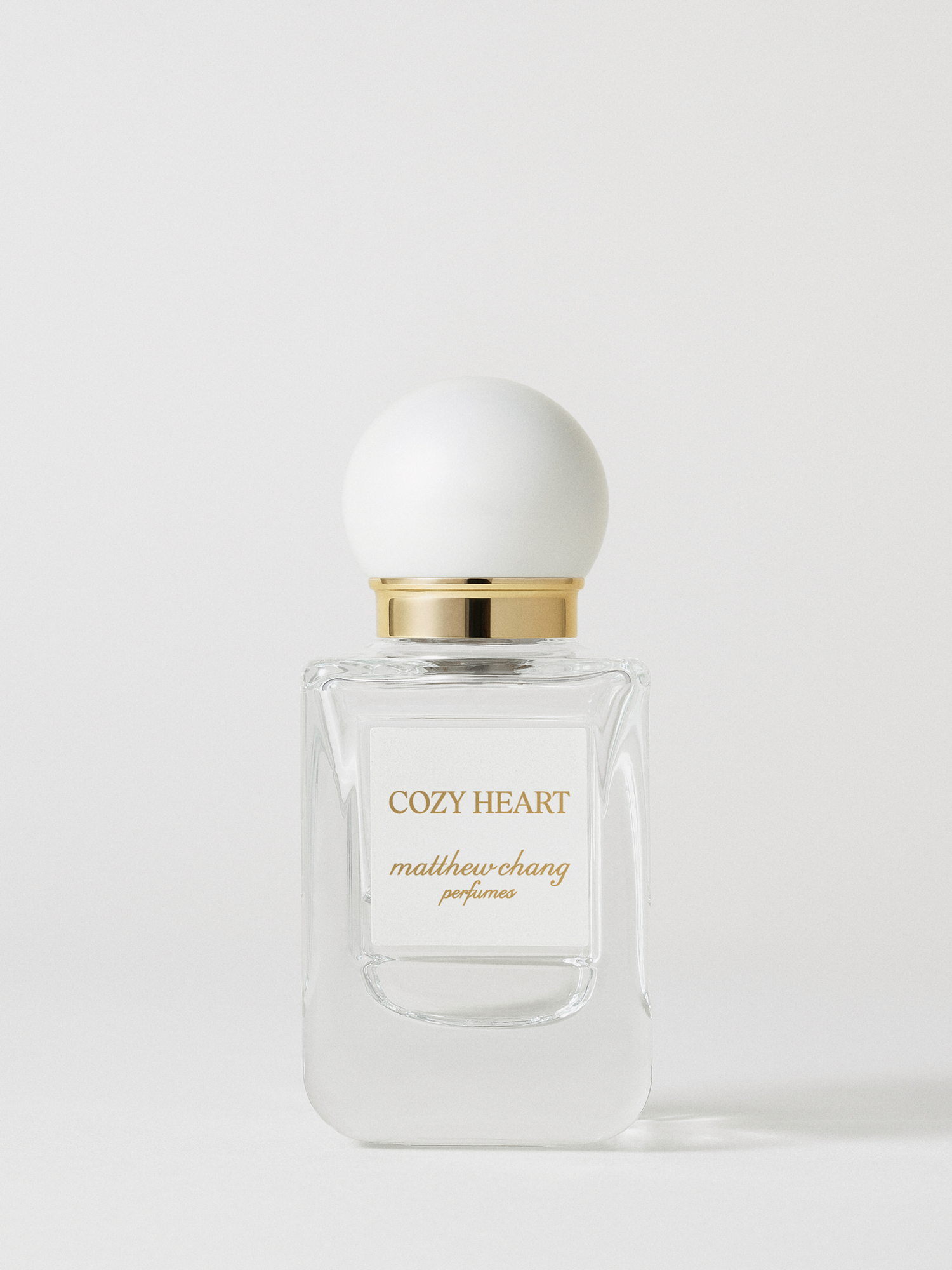 Fotografia prodotto del profumo coreano Cozy Heart firmato Matthew Chang Perfume, presentato in un elegante flacone in vetro trasparente con etichetta frontale, tappo a sfera bianco opaco e collo dorato, su sfondo chiaro minimal che valorizza il design raffinato della fragranza.