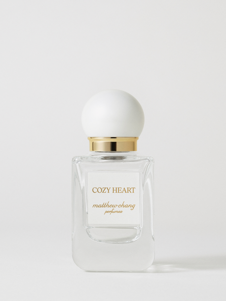 Fotografia prodotto del profumo coreano Cozy Heart firmato Matthew Chang Perfume, presentato in un elegante flacone in vetro trasparente con etichetta frontale, tappo a sfera bianco opaco e collo dorato, su sfondo chiaro minimal che valorizza il design raffinato della fragranza.