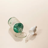 Flacone in vetro verde acqua del AXIS-Y Artichoke Intensive Skin Barrier Ampoule inclinato e aperto, con una goccia di siero che scende sul tappo appoggiato accanto. Sfondo beige chiaro, atmosfera luminosa ed elegante.

