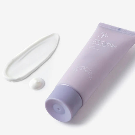 Tubo lilla di crema viso SOROCI Morning Drizzle Calming Cream inclinato su sfondo chiaro, con una goccia di prodotto e una fascia bianca posizionate accanto.