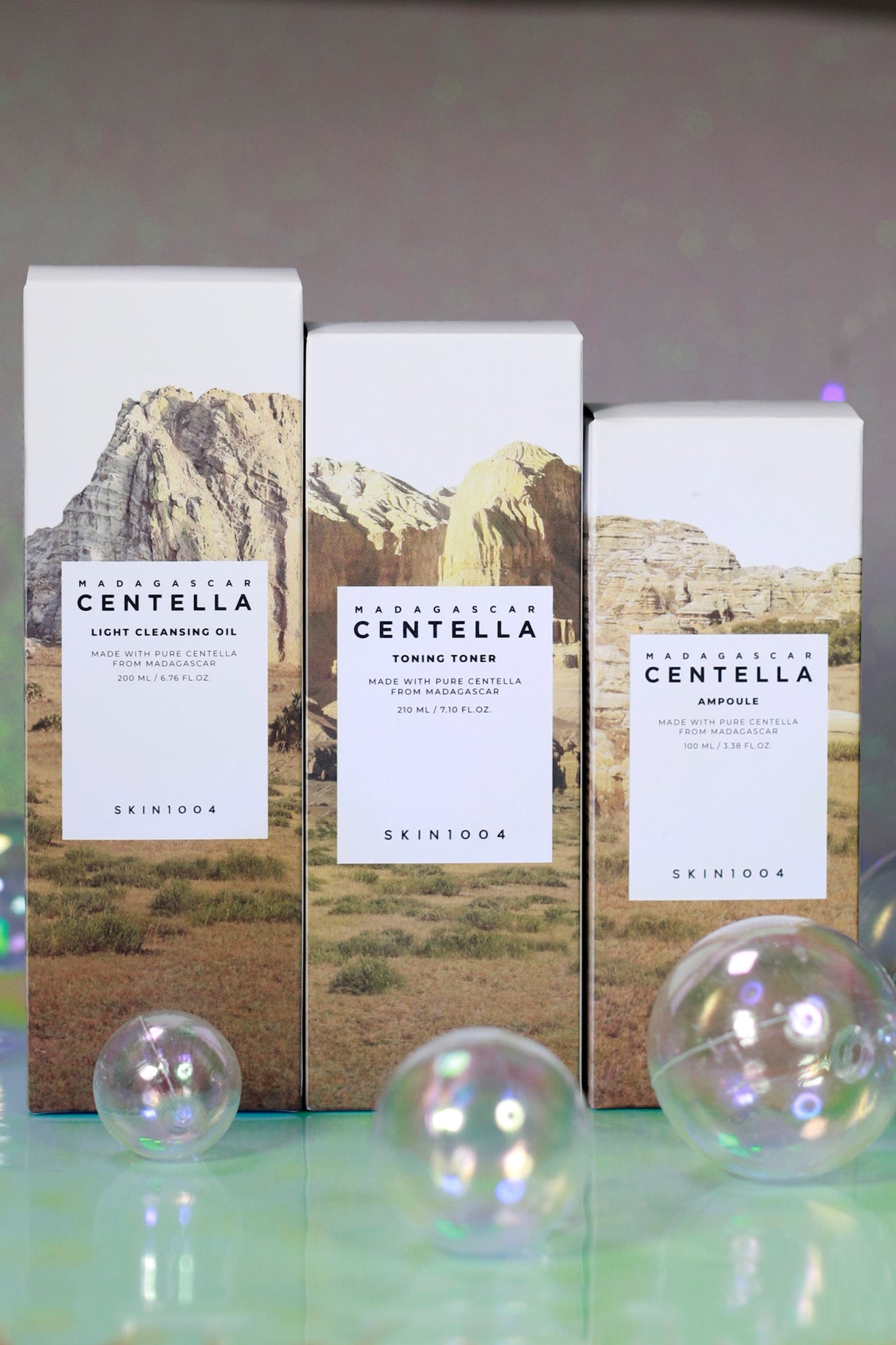Tre confezioni in cartone dei prodotti SKIN1004 Centella disposte in verticale su fondo colorato con bolle trasparenti: da sinistra Light Cleansing Oil, Toning Toner e Ampoule, tutte con paesaggio roccioso stampato sulla confezione.