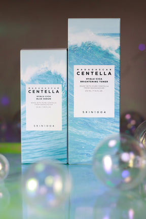 Set skincare Skin1004 Hyalu-Cica con Brightening Toner da 210 ml e Blue Serum da 50 ml, confezioni azzurre con grafica effetto onda, su sfondo riflettente con bolle traslucide.