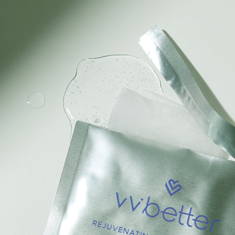 Dettaglio di una maschera viso aperta vvbetter con confezione argentata, liquido trasparente con microbolle versato e tessuto della maschera visibile su sfondo chiaro.