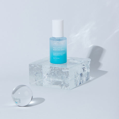 Flacone azzurro del siero viso Wbetter TECA Lifting Moisture da 30ml posizionato su un blocco trasparente tipo ghiaccio, con sfera decorativa su sfondo chiaro