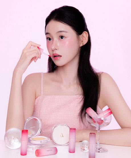 Modella con make-up luminoso che applica una tinta labbra Aperire, circondata da lip tint e cushion su tavolo