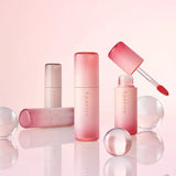 Set di lip tint Aperire con flaconi rosa gradient e applicatore doe-foot aperto, posizionati su sfondo chiaro con sfere in vetro
