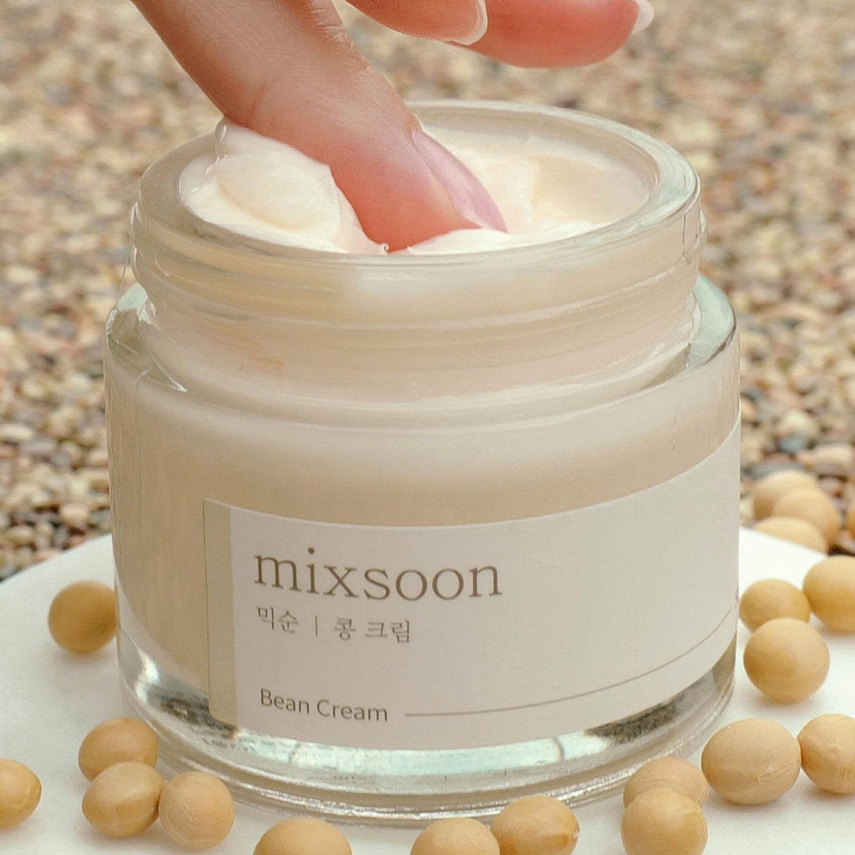 Un dito preleva la crema da un vasetto di Mixsoon Bean Cream aperto, circondato da piccoli fagioli beige su una superficie chiara con sfondo granuloso."
