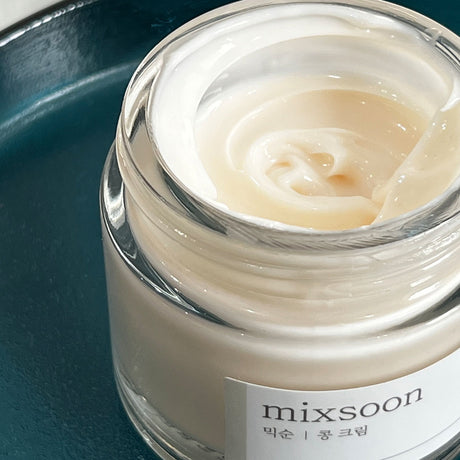 Vasetto aperto della Mixsoon Bean Cream ripreso da vicino, con consistenza cremosa visibile all’interno, appoggiato su superficie blu lucida.
