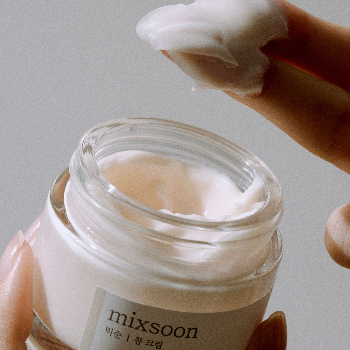 Vasetto aperto di Mixsoon Bean Cream tenuto in mano, con una dita che preleva una generosa quantità di crema dalla texture densa e soffice.
