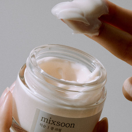 Vasetto aperto di Mixsoon Bean Cream tenuto in mano, con una dita che preleva una generosa quantità di crema dalla texture densa e soffice.

