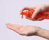 Flacone arancione Cafuné Enhancer Hair Tonic eroga il tonico spray su una mano, mostrando la texture liquida leggera del trattamento lenitivo per cute, ideale contro prurito e forfora.
